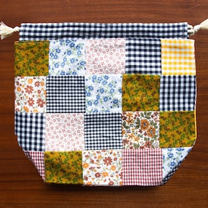 May include: A patchwork drawstring bag with a variety of patterned squares. The bag features squares of floral, gingham, and solid fabrics in colors like blue, yellow, and burgundy. The top edge is black and white gingham with a drawstring closure.