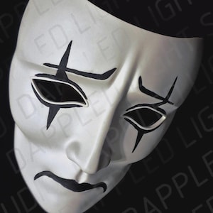 Pode incluir: Uma máscara teatral branca com marcas pretas ao redor dos olhos e da boca. A máscara tem uma forma ligeiramente curvada e uma superfície lisa. As marcas pretas incluem desenhos em forma de cruz acima dos olhos e uma linha curva para a boca.