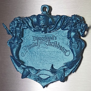 Può includere: Un ornamento in metallo blu turchese a forma di sirena con un teschio di pirata e ossa incrociate al centro. L'ornamento presenta il testo "Disneyland's Pirates of the Caribbean The Original".