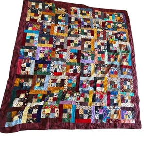 May include: A rectangular patchwork quilt featuring a grid of multi-colored fabric squares in various patterns, including floral, geometric, and solid prints. The quilt is bordered by a solid dark maroon fabric frame on all four sides.