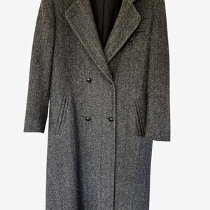 May include: A long, double-breasted coat in a grey and black herringbone pattern. The coat features a notched lapel, two vertical slit pockets, and four black buttons on the front. A label inside the collar reads J.G. HOOK.