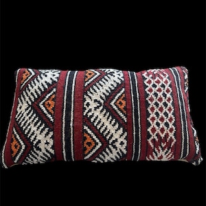 May include: A rectangular throw pillow featuring a woven textile pattern in deep red, black, white, and orange. The design consists of vertical stripes, geometric diamond shapes, and intricate lattice motifs across the entire surface of the pillow.
