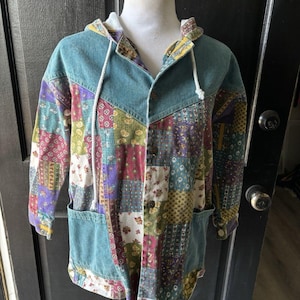 May include: A hooded denim jacket with a patchwork design. The jacket features a teal blue denim hood and pockets, with a colorful patchwork pattern of floral and geometric designs. It has a button-up front and a drawstring hood.