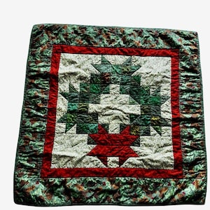 May include: A quilted blanket with a central starburst design in red, green, and white. The starburst is framed by a red border and surrounded by a green patterned edge. The quilt has a handmade appearance.