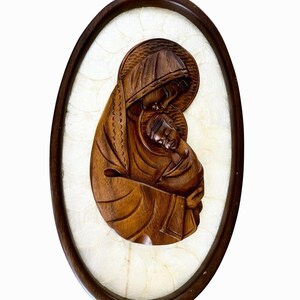 May include: An oval wall plaque featuring a dark brown carved wooden relief of a parent figure holding a child. The carving is set against a textured, off-white, translucent background material and enclosed within a dark brown wooden oval frame.