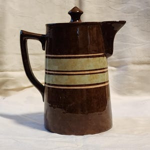 May include: A brown ceramic coffee pot with a lid and a handle. The pot has three bands of green and pink around the middle.