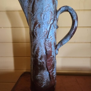 May include: A tall, blue and brown ceramic jug with a unique, textured design. The jug has a wide mouth and a handle that curves up and over the top.