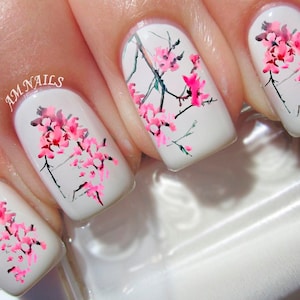 Könnte beinhalten: Weißer Nagellack mit rosa Kirschblüten-Nagelkunst-Aufklebern. Die Nagelkunst zeigt zarte Zweige mit rosa Blüten und grünen Blättern.