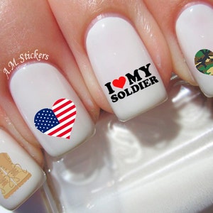 Puede incluir: Cuatro uñas blancas con diseños de nail art. Una uña tiene un corazón rojo con las palabras "I love my soldier" en letras negras. Otra uña tiene un corazón con un diseño de bandera estadounidense. Una tercera uña tiene un corazón con un patrón de camuflaje. La cuarta uña tiene un par de botas marrones.
