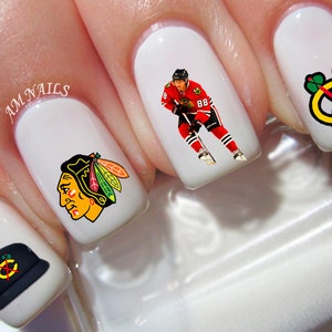 May include: White nail art featuring Chicago Blackhawks hockey team designs. The designs include the team logo, a player in uniform, and a hat with the team logo. The text "AM NAILS" is also visible.