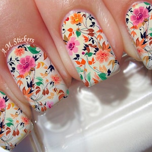 May include: Close-up of four fingernails with floral nail art. The nails are painted with a white base and feature a colorful floral design with pink, orange, and green accents. The text "A.M. Stickers" is visible on one of the nails.