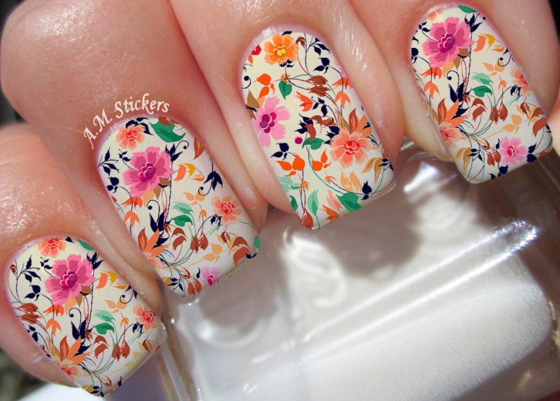 May include: Close-up of four fingernails with floral nail art. The nails are painted with a white base and feature a colorful floral design with pink, orange, and green accents. The text "A.M. Stickers" is visible on one of the nails.