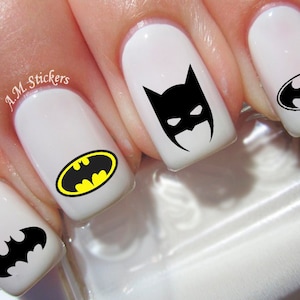 May include: White fingernails with black and yellow Batman logo nail decals. The decals feature different variations of the Batman logo, including the iconic bat symbol and a masked Batman head.