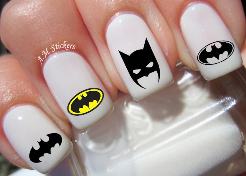 May include: White fingernails with black and yellow Batman logo nail decals. The decals feature different variations of the Batman logo, including the iconic bat symbol and a masked Batman head.