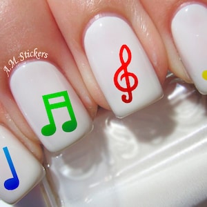 May include: Five fingernails with white nail polish and colorful music note decals. The notes are green, blue, yellow, and red. The red note is a treble clef.