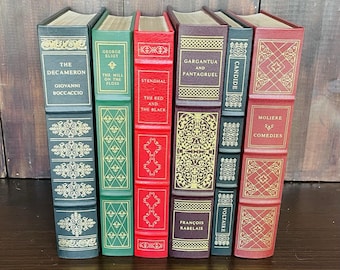 May include: A collection of classic books with decorative covers. Titles include "The Decameron," "The Mill on the Floss," and "Candide." The books are in various colors, including green, red, and black, with gold accents. The books are arranged upright.