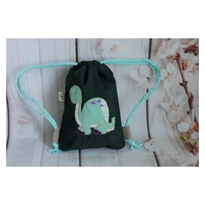 Könnte beinhalten: Eine grüne Kordeltasche mit einem hellgrünen Dinosaurier-Applikationen. Die Tasche hat hellblaue Kordeln.