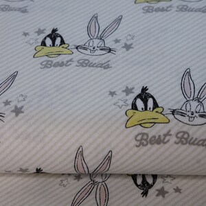 Könnte beinhalten: Weißer Stoff mit einem sich wiederholenden Muster von Bugs Bunny und Daffy Duck Comicfiguren. Die Figuren sind in Schwarzweiß mit gelben Akzenten. Der Stoff hat einen grau-weiß gestreiften Hintergrund mit den Worten "Best Buds", die sich durchgehend wiederholen.