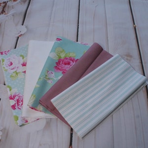 Op de afbeelding: Vier stofstalen in verschillende patronen en kleuren. De stalen omvatten een witte stof, een lichtblauwe stof met witte stippen en roze rozen, een lichtblauwe stof met witte strepen en een lichtroze stof met een bloemenpatroon.