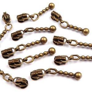 Può includere: Un set di otto tiretti per cerniera in oro antico con una catena e dettagli perlati. Ogni tiretto ha una perla rotonda in metallo all'estremità.