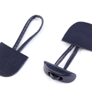 Pode incluir: Dois puxadores de zíper de couro preto com um botão de plástico preto. Os puxadores têm a forma de um retângulo arredondado.