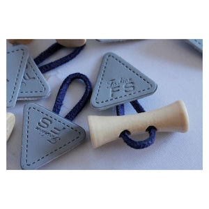 Pode incluir: Etiquetas de couro cinzas em forma de triângulo com o texto "Fashion FS" em relevo. As etiquetas estão presas a um cordão azul e a um botão de madeira.