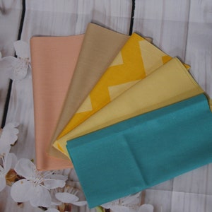 以下が含まれることがあります： 桃色、ベージュ、黄色、ターコイズなど、さまざまな色のファブリック正方形6枚セット。正方形は扇形に配置され、桃色の正方形が上、ターコイズの正方形が下にあります。