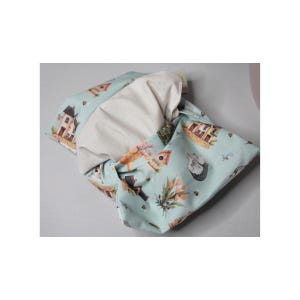 May include: A light blue fabric item featuring a print of houses, animals, and flowers. The item is knotted, showing a cream-coloured interior. The design is soft and inviting, possibly a baby or nursery item. The item is likely made of cotton.