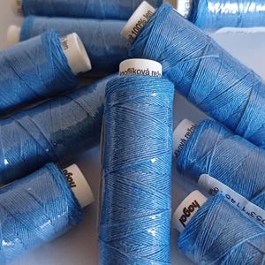 May include: A close-up of several spools of blue thread. The spools are labelled with the text "hagal" and "flíková rež".