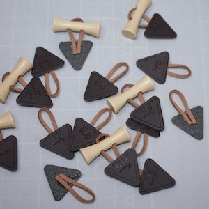 Könnte beinhalten: Eine Sammlung brauner Leder-Dreiecksanhänger mit einem kleinen Holz-Toggle und einem braunen Lederband. Die Anhänger haben den Text "The Feel" geprägt.