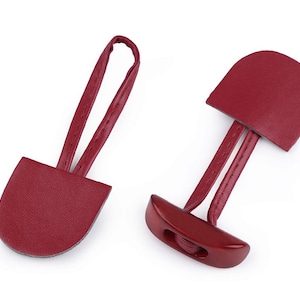 Könnte beinhalten: Zwei rote Leder-Knöpfe mit einer Schlaufe zum Befestigen von Kleidung. Die Knöpfe haben eine abgerundete Form und sind durch ein Lederband verbunden.