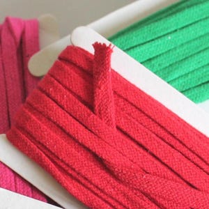 May include: Three spools of flat fabric cord in vibrant colours, including a bright pink, a bold red, and a vivid green. Each cord is neatly wound around a white rectangular cardboard holder, showcasing a consistent texture and width.