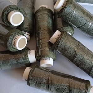 May include: A close-up of several spools of dark green thread. The spools are wrapped in clear plastic and have white labels with text in various languages. The text on the labels includes "80 m TEX 40", "Knopflik", and "Knopfliko".