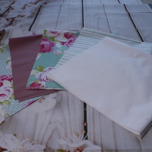 Könnte beinhalten: Vier Stoffmuster, zwei mit Blumenmustern, eines mit Streifen und eines einfarbig weiß. Die Blumenmuster sind auf hellblauem Grund mit rosa Rosen und weißen Punkten. Der gestreifte Stoff ist hellblau und weiß. Der einfarbige Stoff ist weiß.