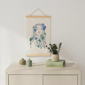 Può includere: Un'opera d'arte da parete con un disegno a tratto di una donna con fiori tra i capelli. L'opera è esposta su una cornice di legno. Una piccola pianta in vaso e dei libri sono su un mobile color crema. Il muro è bianco.
