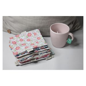 May include: A pile of floral fabric squares with pink and red flowers, beside a pink mug with a teabag. The fabric squares have small tags attached. A grey cushion is in the background.
