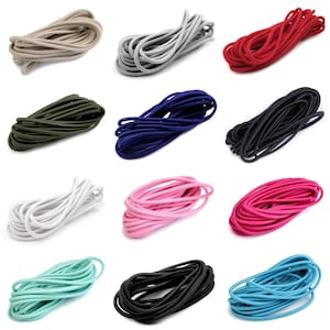 Op de afbeelding: Assortiment van 12 spoelen elastisch koord in verschillende kleuren, waaronder zwart, wit, rood, roze, blauw, groen en beige. Het elastische koord is opgerold en in rijen gerangschikt.