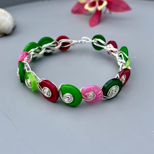 May include: A handmade bracelet featuring colorful, round beads in shades of green, pink, and red, strung together with silver wire. The beads have a spiral design. The bracelet is displayed on a gray surface, with a flower in the background.