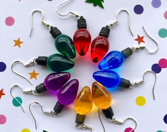 May include: A collection of eight dangle earrings shaped like miniature translucent Christmas light bulbs in red, green, blue, purple, and orange. Each bulb features a black plastic base and a silver-toned metal hook for ear attachment.