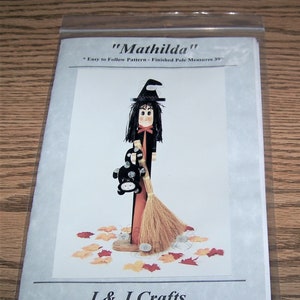 May include: A craft pattern for a witch doll named "Mathilda". The pattern includes instructions for making a witch doll with a black dress, a black hat, and a broom. The doll is holding a black and white cat. The pattern is from J & J Crafts, 203 4th Street, P.O. Box 133, Grafton, IA 50440, 515-748-2748.