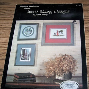 May include: A leaflet titled "Award Winning Designs" by Judith Sandy, featuring three framed cross-stitch patterns. The leaflet is from Graphique Needle Arts and costs $2.00. The leaflet is labeled "Leaflet #2".