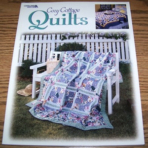 May include: A book titled "Cozy Cottage Quilts" by Leisure Arts. The cover features a quilt with floral patterns on a white bench in a garden setting. The book also shows a quilt with a geometric pattern on a bed.