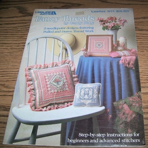 May include: A book cover for a needlepoint project titled "Fancy Threads" by Candy Kirby. The cover features two needlepoint pillows, one with a floral design and the other with a monogram. The book includes step-by-step instructions for beginners and advanced stitchers.