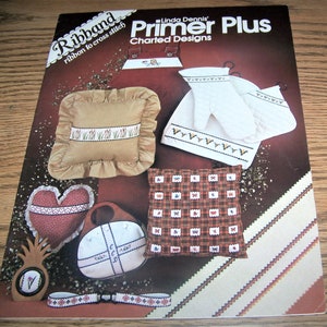 May include: A book titled "Ribbon Primer Plus Charted Designs" by Linda Dennis. The book features various ribbon embroidery projects, including a pillow, a heart, a purse, and oven mitts.