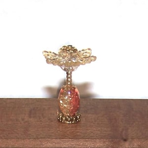 May include: A decorative bottle stopper with a flower-shaped top and a stem. The stopper has a gold-colored flower design and a red and gold-colored base. The stopper is sitting on a brown surface.