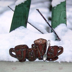 May include: A brown ceramic coffee-themed pin with the letters "JAVA" and a small silver spoon. The pin features four coffee cups and is set against a snowy background with green fence posts.