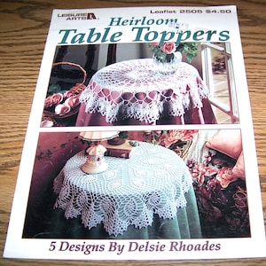 May include: A crochet pattern booklet titled "Heirloom Table Toppers" by Leisure Arts. The booklet features two photos of crocheted tablecloths, one on a pink tablecloth and one on a green tablecloth. The booklet is for sale for $4.50 and is leaflet number 2505. The text "5 Designs By Delsie Rhoades" is printed at the bottom of the booklet.