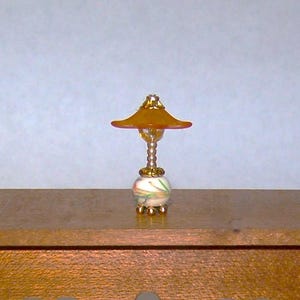 May include: Miniature lamp with a golden-yellow lampshade and a decorative base. The base is white with colorful swirls and green accents. The lamp has a gold-colored stem and base, and is designed for a dollhouse or miniature setting.