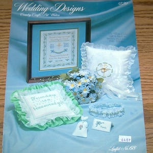 May include: A craft pattern booklet titled Wedding Designs featuring a framed cross-stitch sampler, a white ring bearer pillow with lace trim, a light green ruffled pillow with embroidered text, a bouquet of blue and white flowers, and a garter.