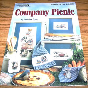 May include: A craft leaflet titled "Company Picnic" by Sandi Gore Evans. The cover features cross-stitch designs of animals at a picnic, including cows, rabbits, and a cat. The designs are framed and accompanied by a pie, carrots, and a bluebird.